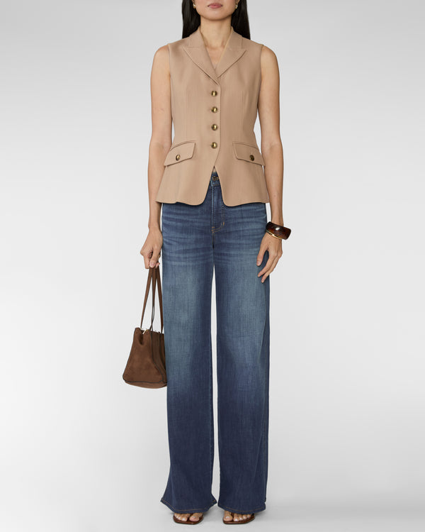 A model wearing a beige sleeveless button-front vest with flap pockets, paired with wide-leg blue jeans, holding a brown handbag against a light gray studio background.