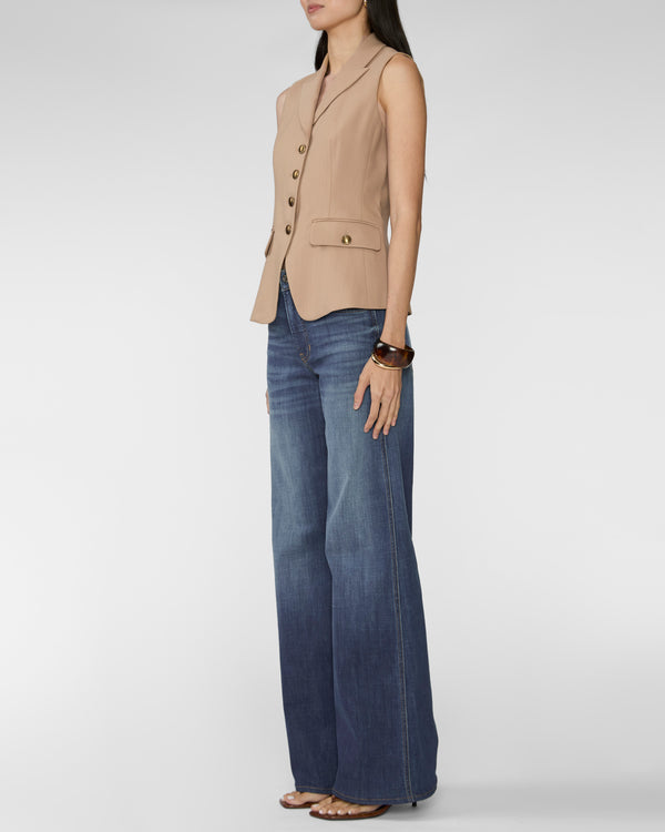 A woman modeling a sleeveless beige vest with gold-tone buttons and flap pockets, paired with wide-leg blue jeans and flat sandals, against a light gray studio background.