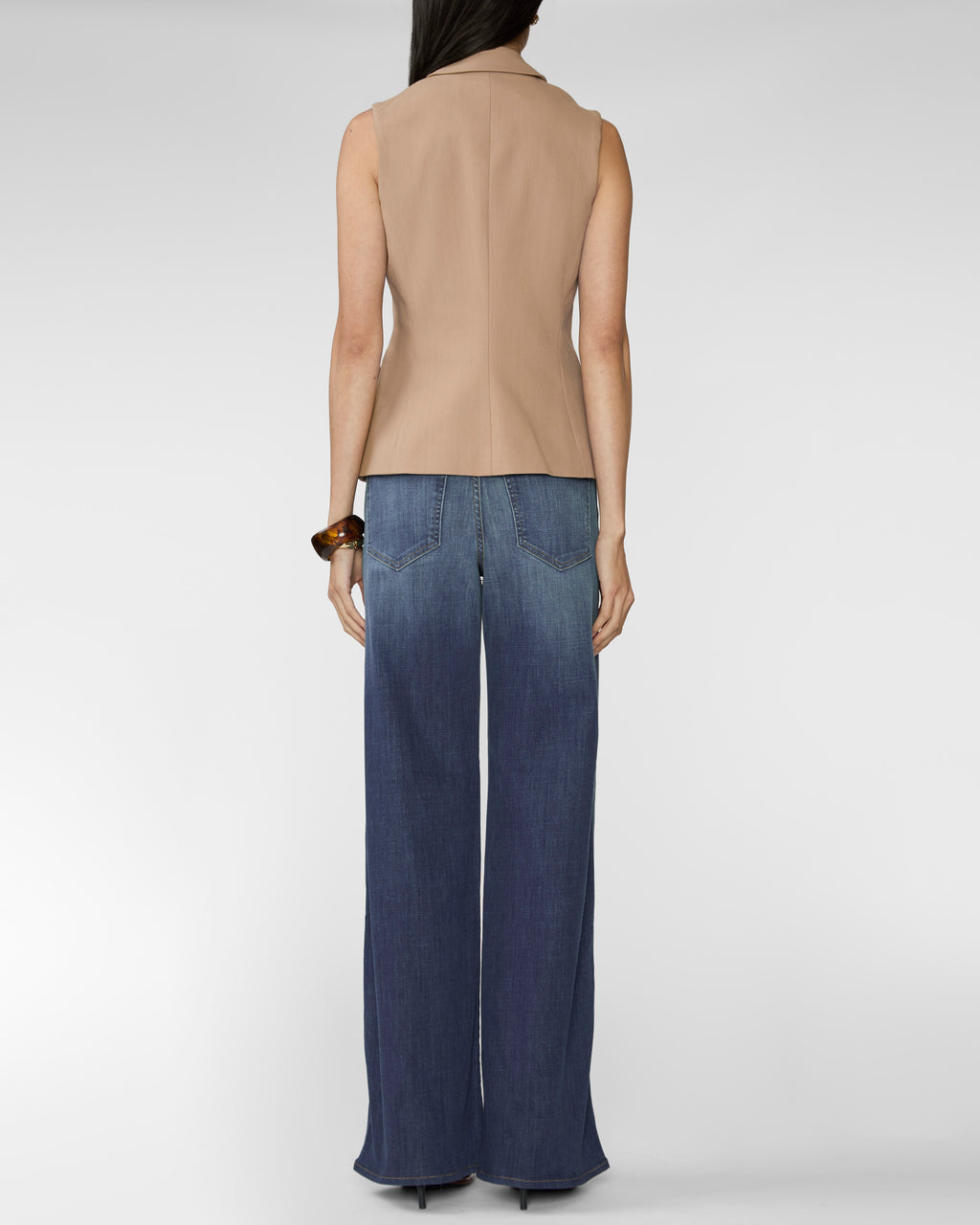 A model shown from the back wearing a sleeveless beige top and wide-leg blue jeans against a light gray studio background.