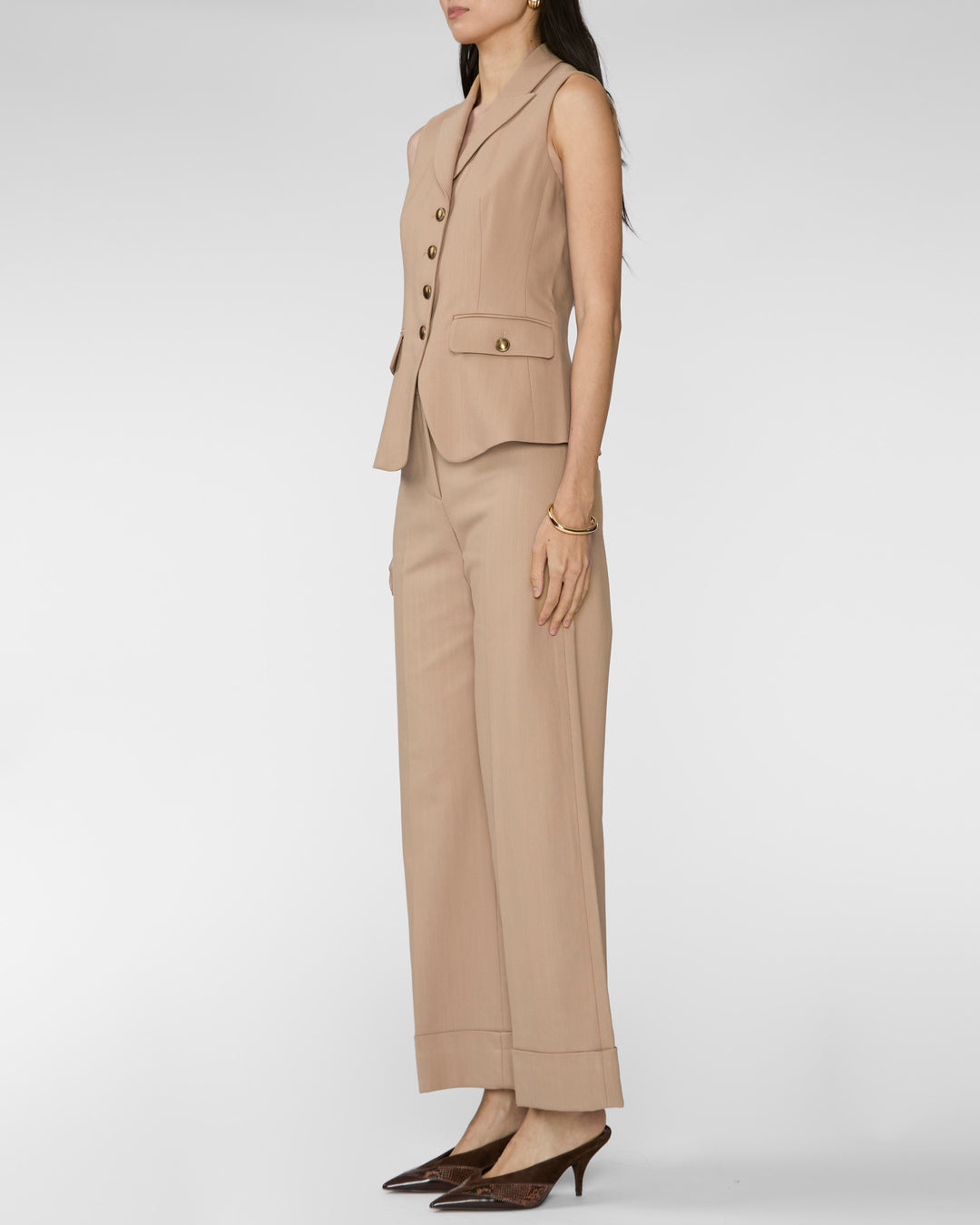 A woman modeling a beige sleeveless blazer and matching wide-leg trousers set, paired with dark brown pointed-toe heels, against a light gray studio background.