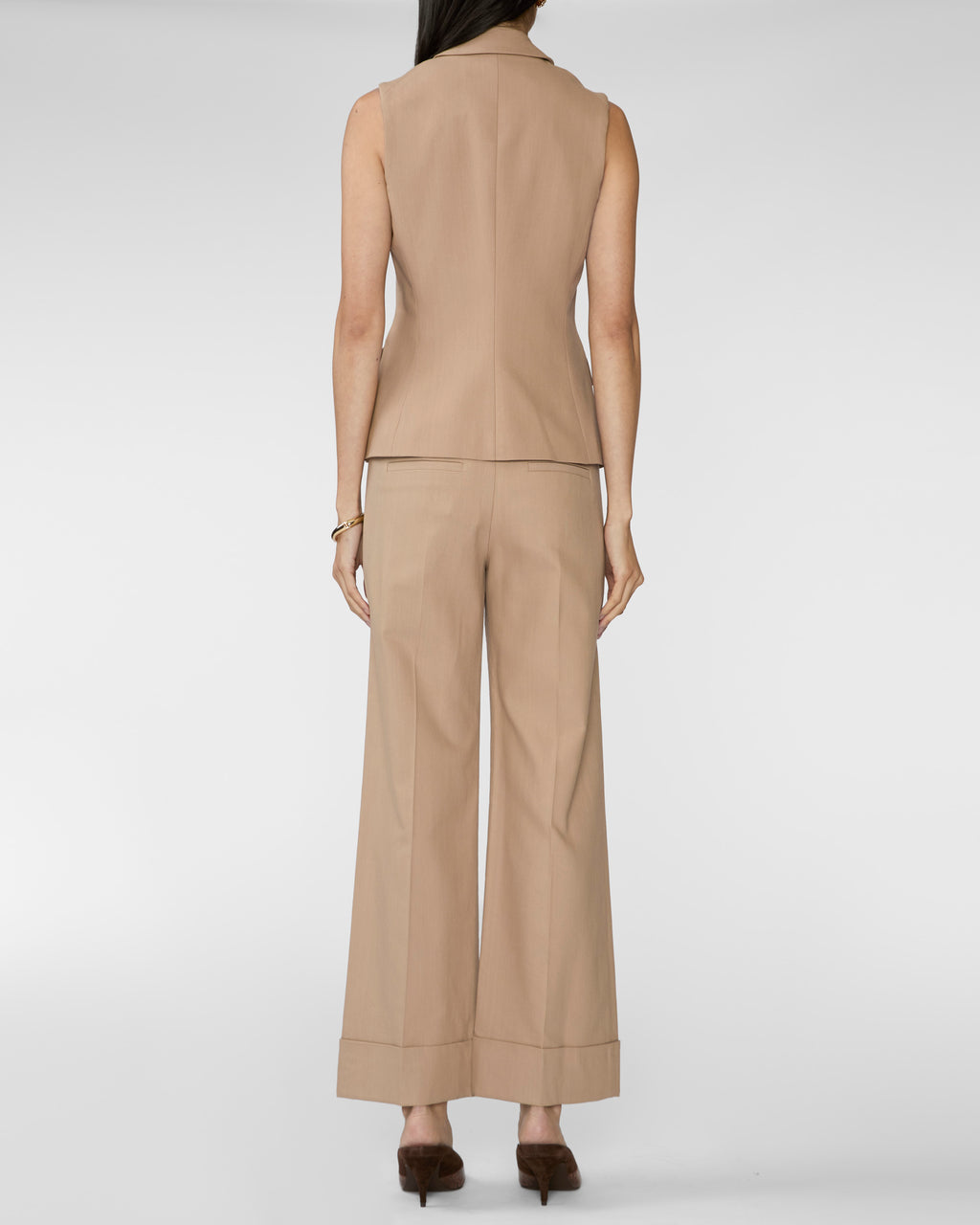A model shown from the back wearing a beige sleeveless vest and matching wide-leg trousers, paired with dark brown pointed-toe heels against a light gray studio background.