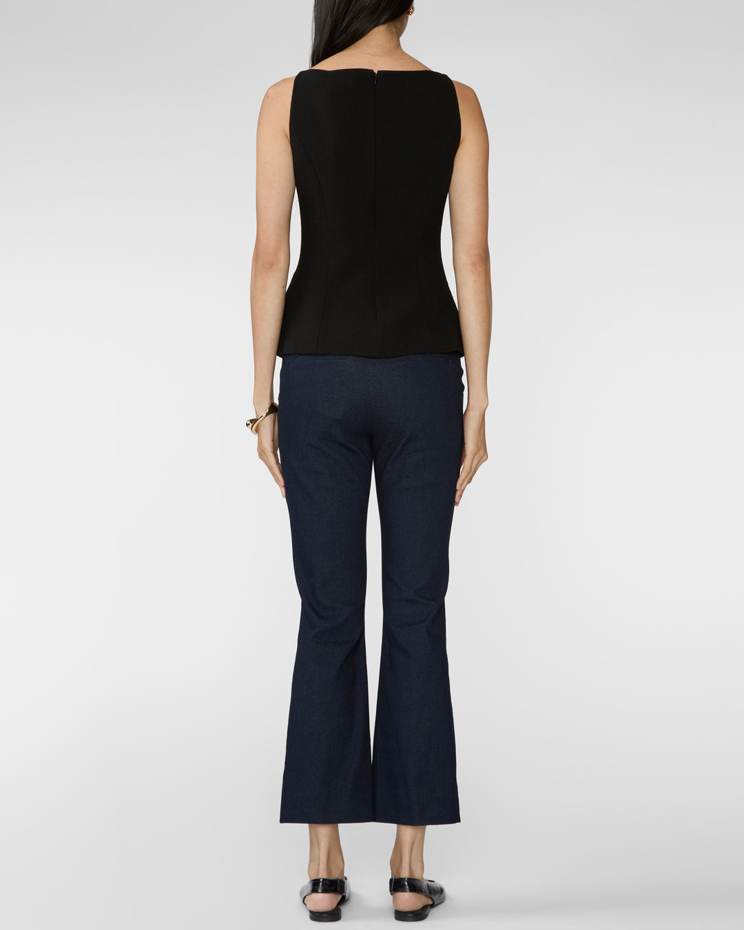 A woman shown from the back wearing a sleeveless black top and dark navy flared pants, standing against a light gray studio background.