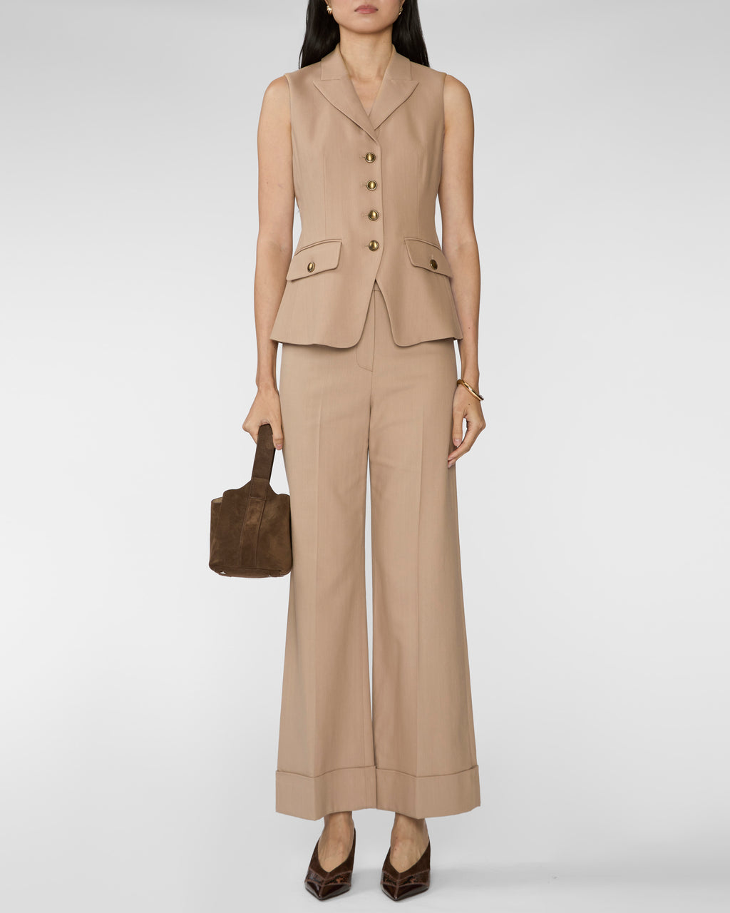 A model wearing a beige sleeveless blazer vest and matching wide-leg trousers, holding a brown handbag against a light gray studio background.