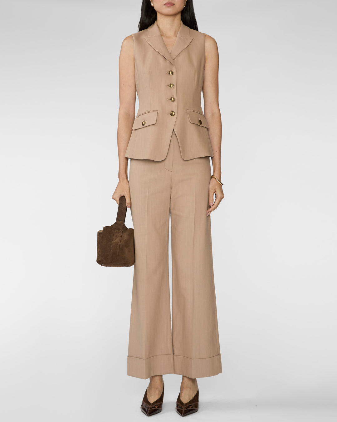 A model wearing a beige sleeveless blazer vest and matching wide-leg trousers, holding a brown handbag against a light gray studio background.