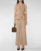 A model wearing a beige sleeveless blazer vest and matching wide-leg trousers, holding a brown handbag against a light gray studio background.