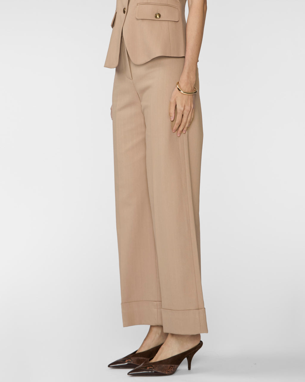 A model is shown from the waist down wearing a matching beige tailored set consisting of wide-leg cropped trousers and a cropped blazer, paired with dark brown pointed-toe heels against a light gray studio background.