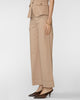 A model is shown from the waist down wearing a matching beige tailored set consisting of wide-leg cropped trousers and a cropped blazer, paired with dark brown pointed-toe heels against a light gray studio background.