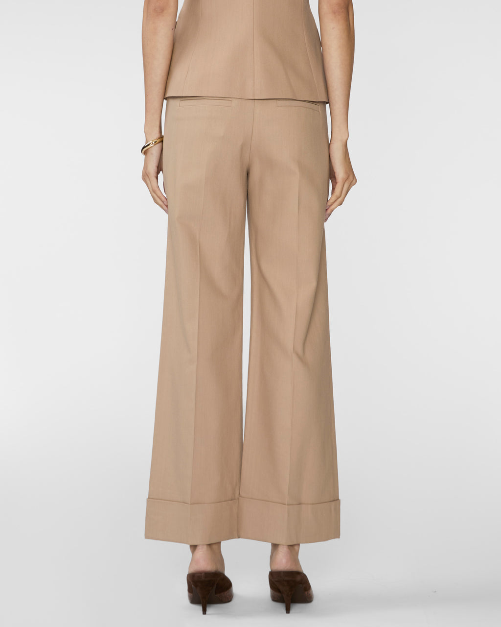 A model shown from the back wearing beige wide-leg cropped trousers, a matching beige top, and dark brown pointed-toe heels against a light gray studio background.