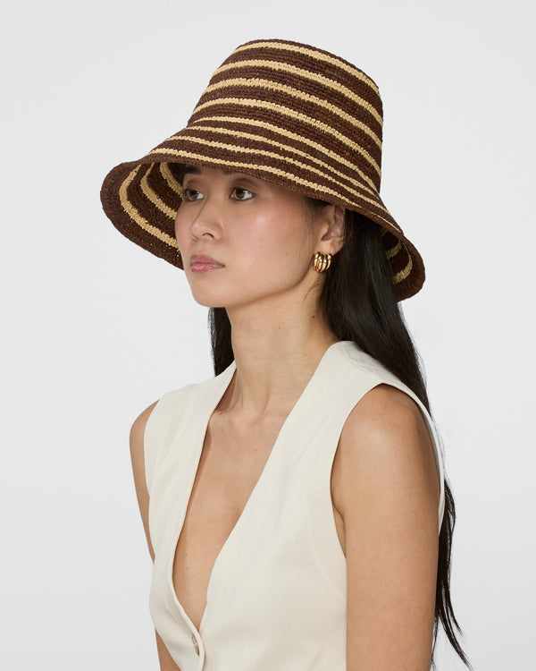 A woman wearing a brown and beige horizontally striped woven bucket hat, paired with a cream-colored sleeveless top, photographed against a solid light gray background.