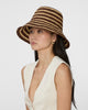 A woman wearing a brown and beige horizontally striped woven bucket hat, paired with a cream-colored sleeveless top, photographed against a solid light gray background.