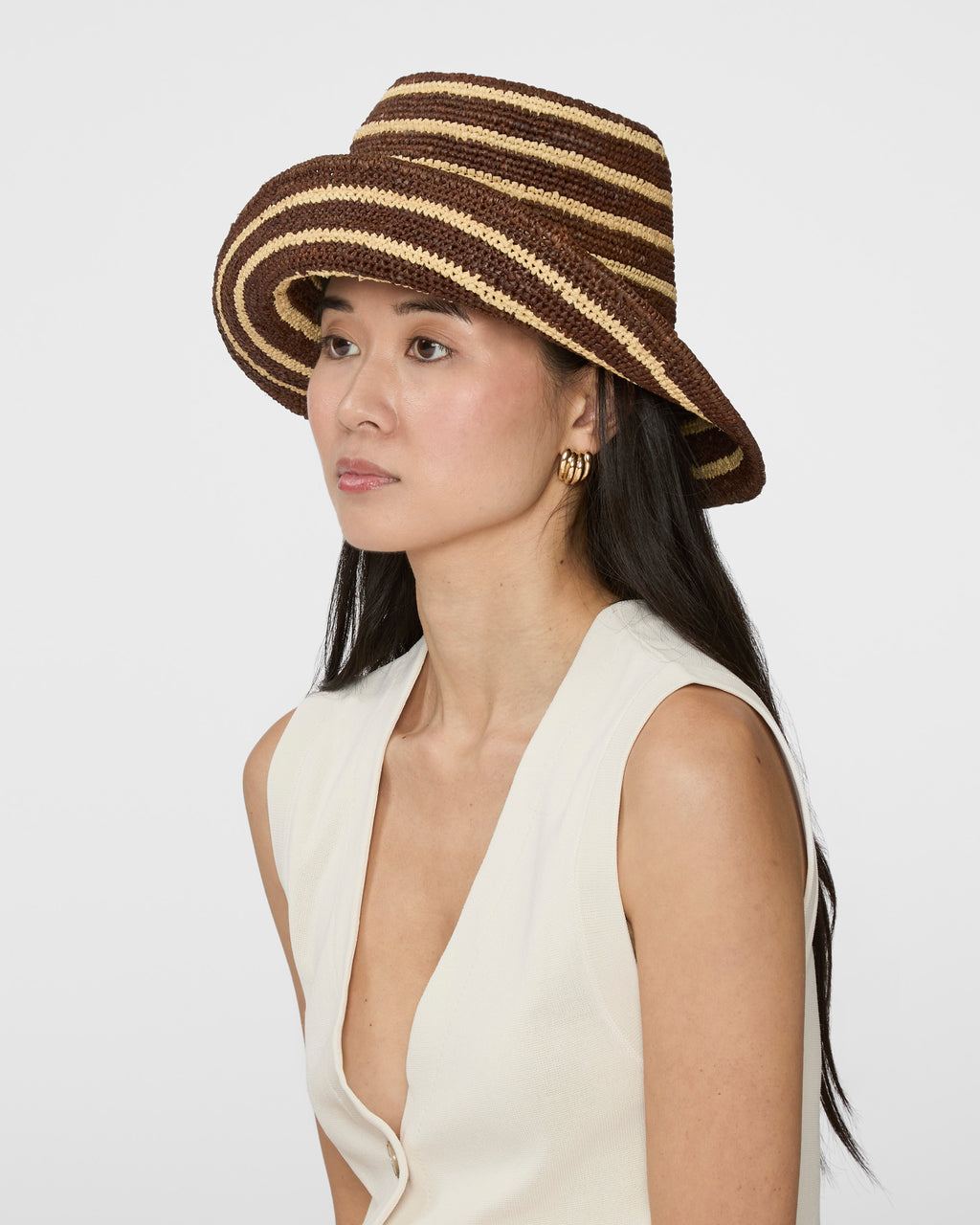 A woman wearing a brown and beige striped woven straw hat, paired with a cream-colored sleeveless top, photographed against a solid light gray background.
