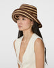 A woman wearing a brown and beige striped woven straw hat, paired with a cream-colored sleeveless top, photographed against a solid light gray background.