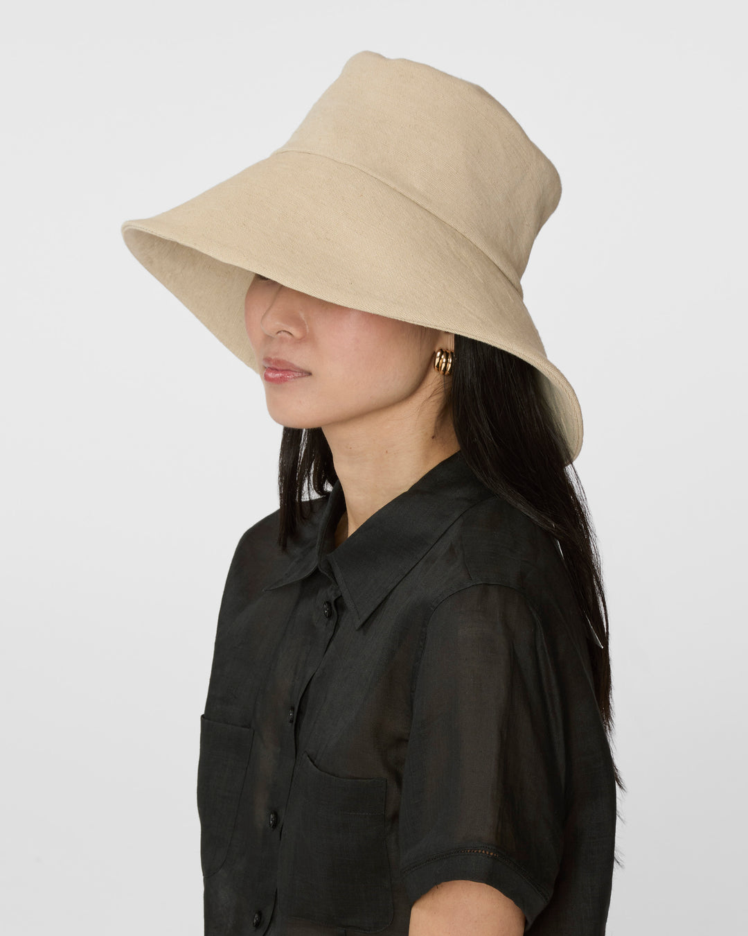 A woman in profile wearing a beige wide-brimmed sun hat and a black short-sleeve button-up shirt, photographed against a light gray studio background.