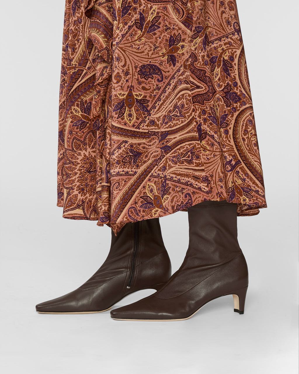 A close-up of a person wearing dark brown leather pointed-toe ankle boots with low kitten heels, paired with a rust-brown and purple paisley patterned skirt, against a light gray studio background.
