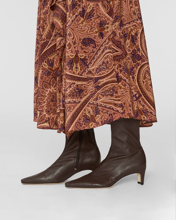 A close-up of a person wearing dark brown leather pointed-toe ankle boots with low kitten heels, paired with a rust-brown and purple paisley patterned skirt, against a light gray studio background.