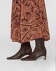 A close-up of a person wearing dark brown leather pointed-toe ankle boots with low kitten heels, paired with a rust-brown and purple paisley patterned skirt, against a light gray studio background.
