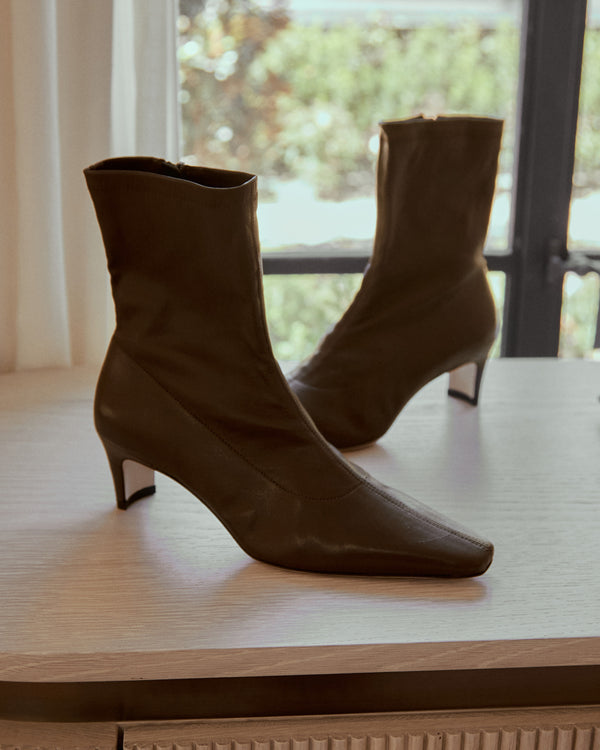 A pair of dark brown leather ankle boots with kitten heels, displayed on a light wood table in front of a window.