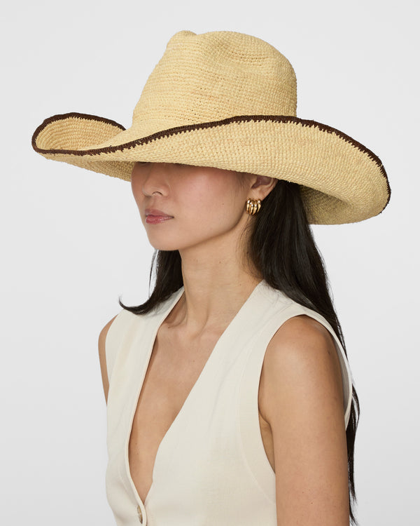 A woman wearing a wide-brimmed straw cowboy hat with a dark brown stitched trim and a cream sleeveless top, photographed against a light gray studio background.