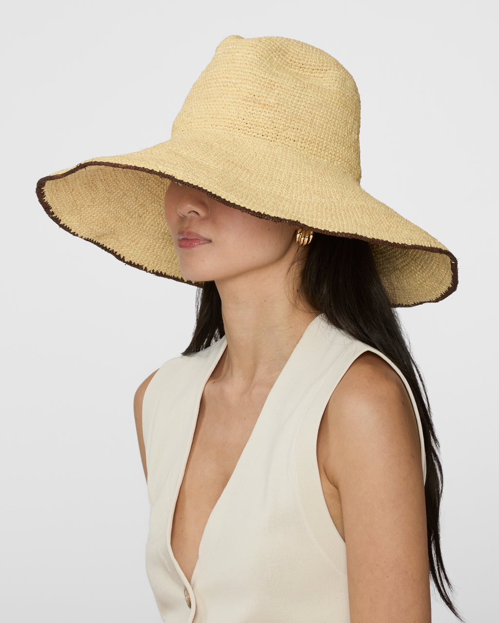 A woman wearing a wide-brimmed straw sun hat in a light beige color with a dark brown frayed edge, paired with a cream-colored sleeveless top, against a plain light gray background.